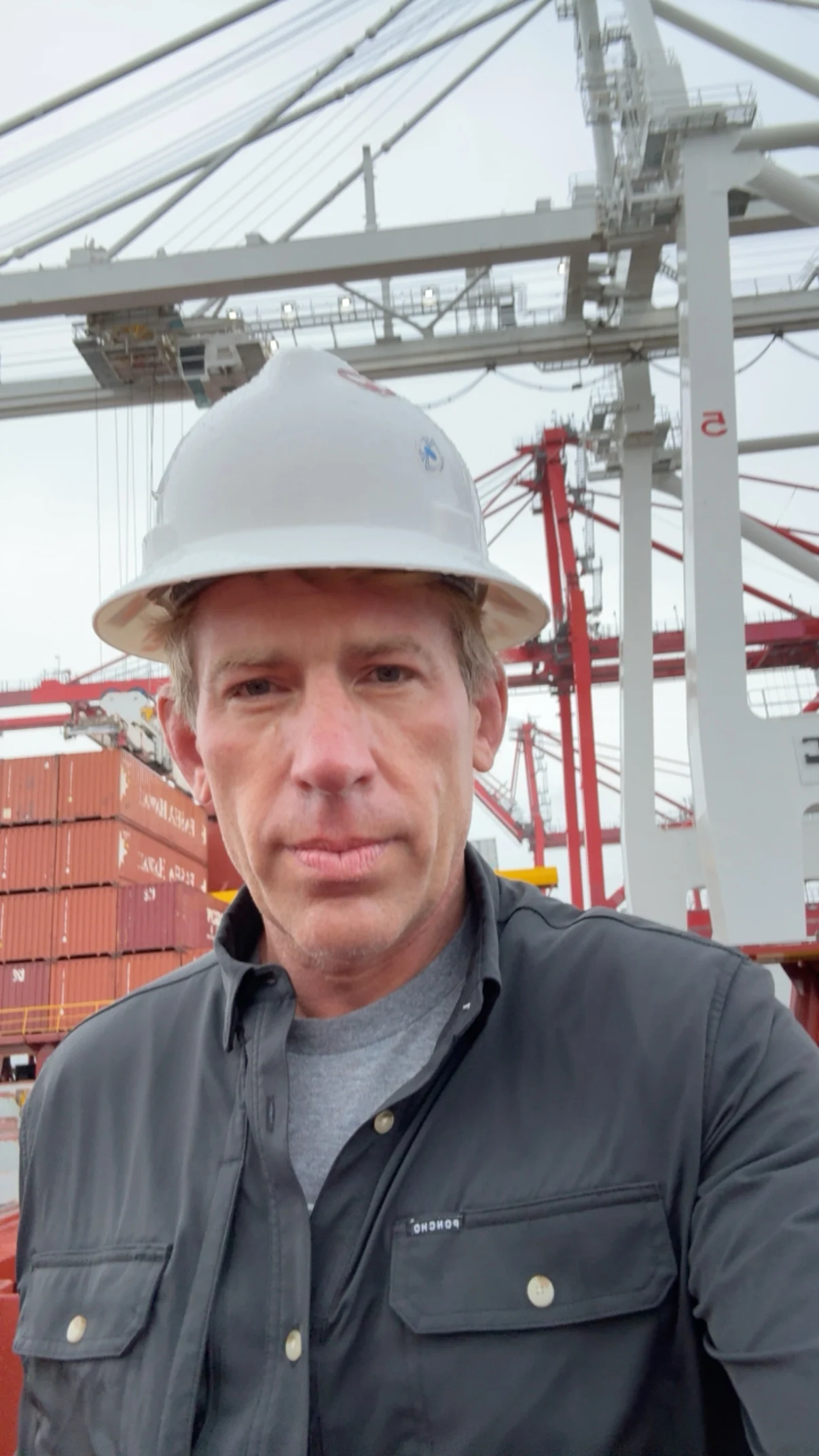 Brian Beckcom in a hard hat during a cargo-ship inspection, with containers and cranes behind him