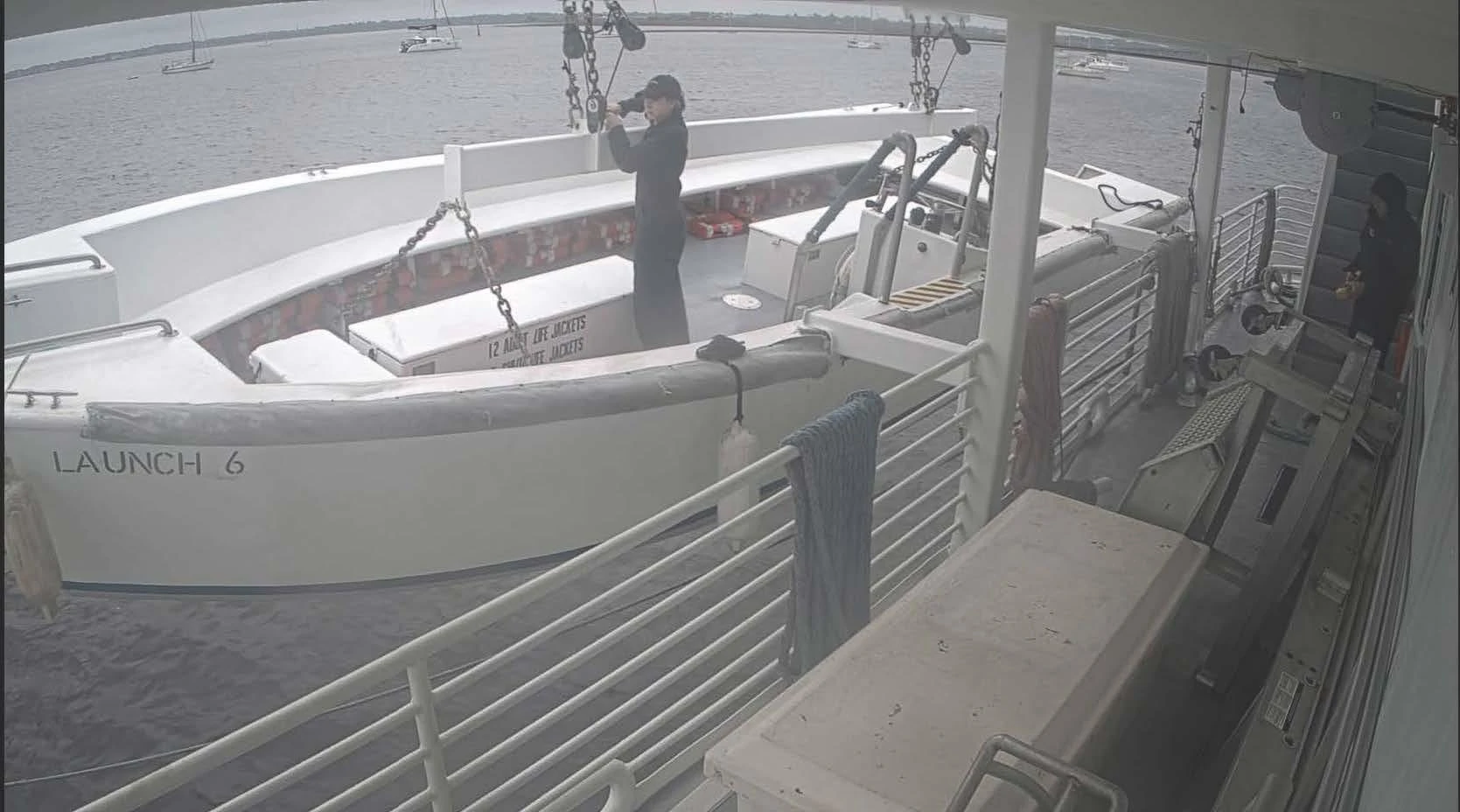 Crew member inside a suspended launch beside a cruise vessel, with lifting chains overhead.