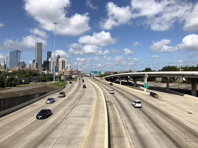 Traffic on Interstate 10 in Houston with downtown buildings to the left and an elevated ramp to the right.