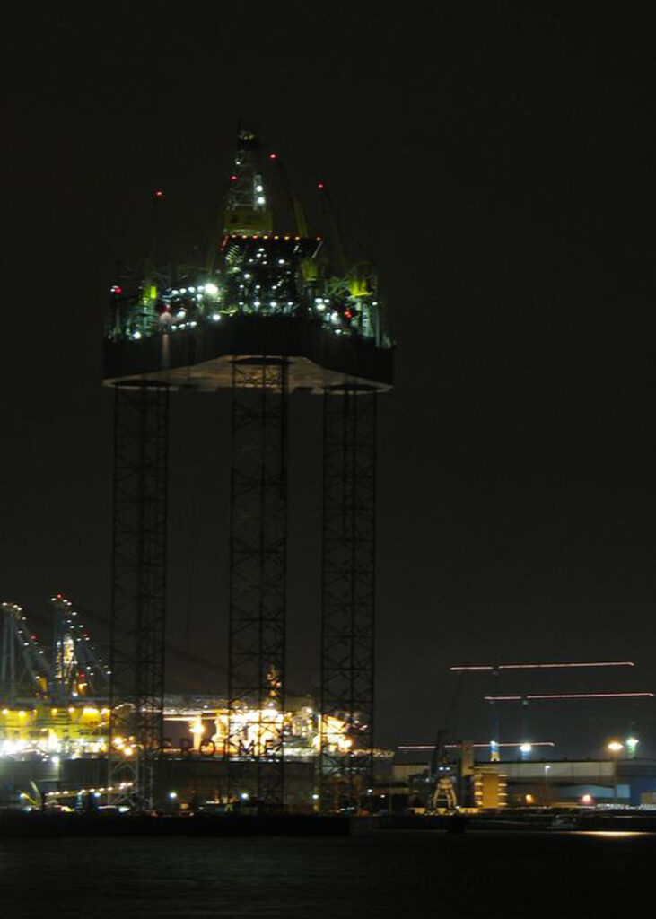 A jack-up drilling rig at night, its hull raised on three lattice legs above the water, deck lights on.