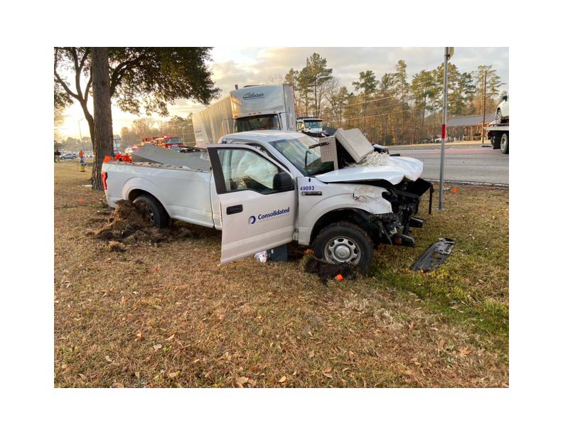 Damaged white Ford F-150 beside the road, with its driver door open and a commercial truck behind it.