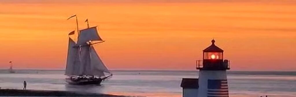 A sailing vessel on the water near a lighthouse beneath an orange sunset sky.